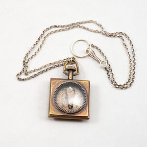 May include: A vintage-style pendant necklace featuring a square, bronze-toned pendant with a clear, circular center. The center showcases a small, light-colored object. The pendant hangs from a silver-colored chain.
