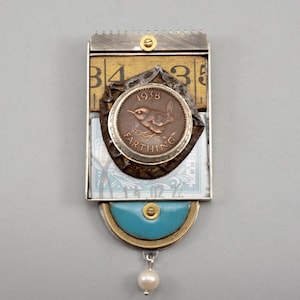 May include: A rectangular brooch featuring a 1938 farthing coin with a bird design, layered over a yellow measuring tape segment and a blue postage stamp. The piece includes a turquoise semi-circle accent, a small white pearl charm, and metal hardware.