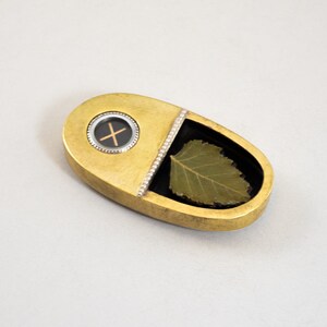 K&ouml;nnte beinhalten: Ein ovaler Anh&auml;nger aus Gold mit einem schwarzen Kreis, der ein Kreuz und ein gr&uuml;nes Blatt in einem transparenten Harzfach enth&auml;lt.