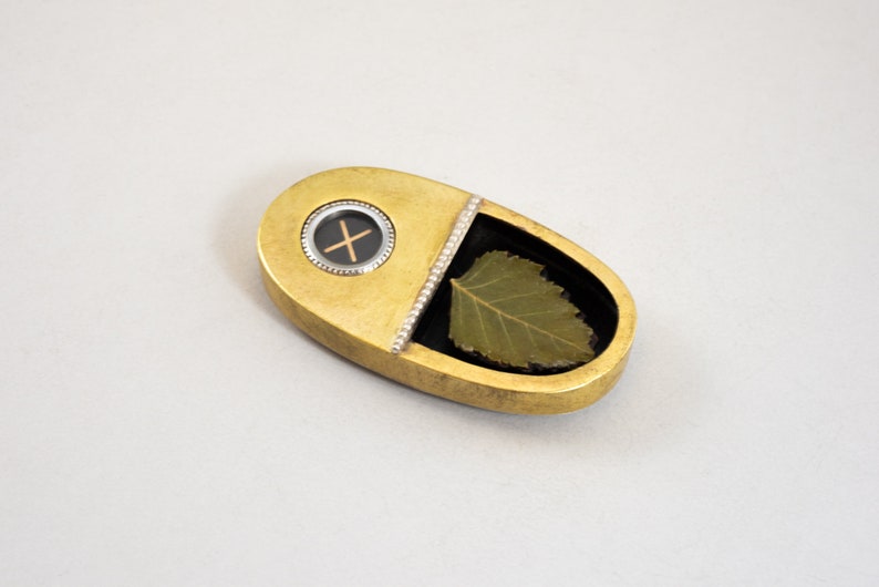 K&ouml;nnte beinhalten: Ein ovaler Anh&auml;nger aus Gold mit einem schwarzen Kreis, der ein Kreuz und ein gr&uuml;nes Blatt in einem transparenten Harzfach enth&auml;lt.