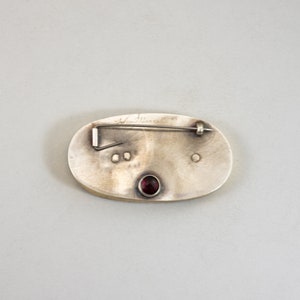 K&ouml;nnte beinhalten: Die R&uuml;ckseite einer ovalen Brosche aus Silber mit einem roten Edelstein in der Mitte. Die Brosche hat eine Nadel und einen Verschluss auf der R&uuml;ckseite und ist mit "Ann Maxwell" signiert.