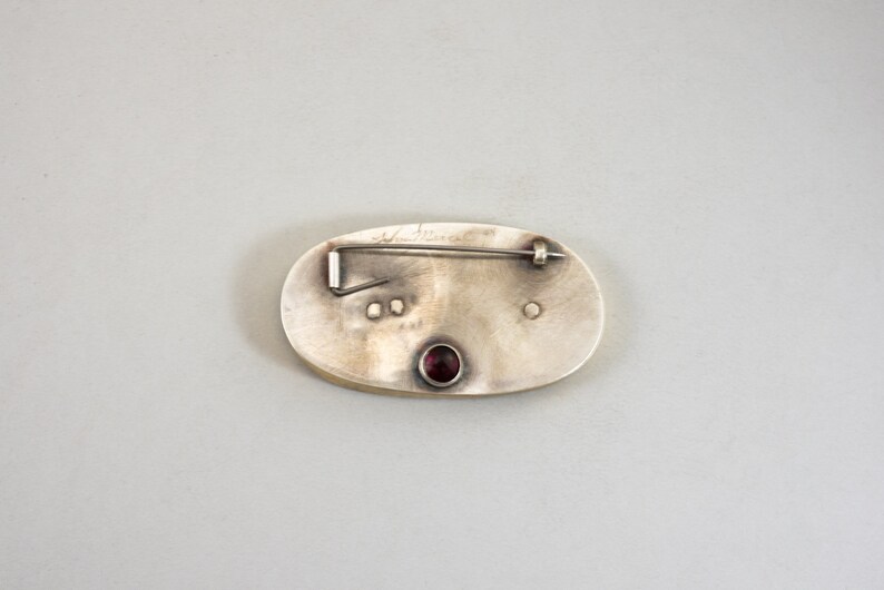 K&ouml;nnte beinhalten: Die R&uuml;ckseite einer ovalen Brosche aus Silber mit einem roten Edelstein in der Mitte. Die Brosche hat eine Nadel und einen Verschluss auf der R&uuml;ckseite und ist mit "Ann Maxwell" signiert.