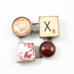 Könnte beinhalten: Quadratische Brosche mit vier Elementen: eine Farthing-Münze von 1954, ein Holz-Scrabble-Stein mit "X8", ein Stück rot-weiß gemusterte Keramik und ein runder, dunkelroter Stein. Die Elemente sind durch silberfarbenes Metall verbunden.