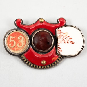May include: A red, white and gold metal brooch with a circular design. The brooch features a red enamel piece with a gear-like design, a round red and white piece with the number "53" on it, a round red stone, and a white porcelain piece with a floral design.