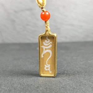 Pode incluir: Um pingente retangular dourado com escrita branca. O pingente é preso a um cordão dourado com uma pequena conta laranja. O pingente tem um design detalhado e provavelmente é uma joia.