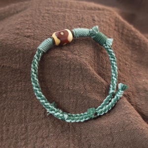 May include: A bracelet constructed from braided sage green cord featuring a single cylindrical bead with a brown and cream-colored pattern. The cord ends are knotted and frayed, and the band is secured with wrapped green thread accents.