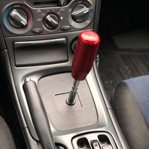 May include: A red aftermarket shift knob for a car. The knob is made of a smooth, red material and has a black metal base. The knob is installed in a car's center console.