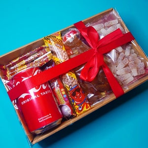 May include: A brown cardboard gift basket filled with a variety of sweets and a can of fizzy drink. The basket is tied with a red ribbon and bow. The sweets include gummy sweets, chocolate sweets, and hard sweets. The can of fizzy drink is red and has the words "Original Taste" and "Delicious & Refreshing" printed on it.