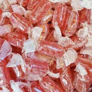 May include: A close-up shot of a pile of red hard candies individually wrapped in clear cellophane. The wrappers have the words "Quality Confectionery" printed on them. The candies are a vibrant red color.