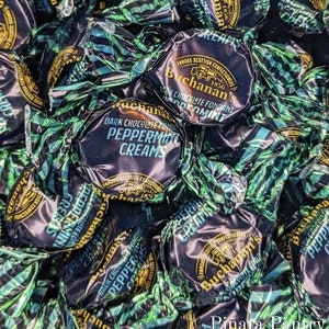May include: A close-up of a pile of individually wrapped dark chocolate peppermint creams. The wrappers are green and black with gold lettering and feature the brand name "Buchanan's" and the text "Famous Scottish Confectioners 1836".