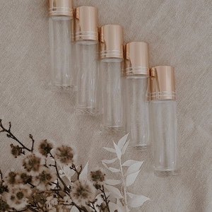 May include: Five clear glass roller bottles with gold lids. The bottles are arranged in a row on a beige fabric background.