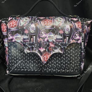 May include: Black satchel bag with a flap featuring a crescent moon pattern. The main body has a print of gothic-themed objects like potion bottles, books, and roses. It has a top handle and a long shoulder strap.
