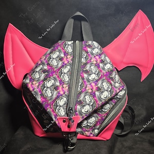 May include: A backpack featuring large, bright pink bat wings on the sides. The main body has a repeating pattern of stylized bat faces against a purple and black galaxy-like background. It includes a black zipper, a top handle, and a small charm.