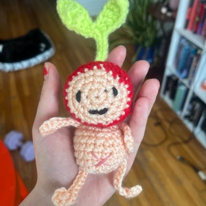 May include: A crocheted amigurumi toy shaped like a person with a red head and green leaves on top. The toy has a pink body and arms and legs.