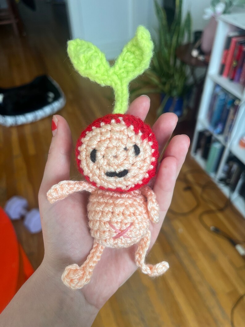May include: A crocheted amigurumi toy shaped like a person with a red head and green leaves on top. The toy has a pink body and arms and legs.