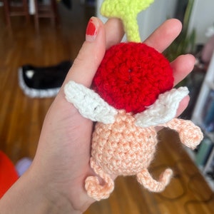 May include: A crocheted plush toy shaped like a cherry with a green stem and white wings. The cherry is red and the body is peach.