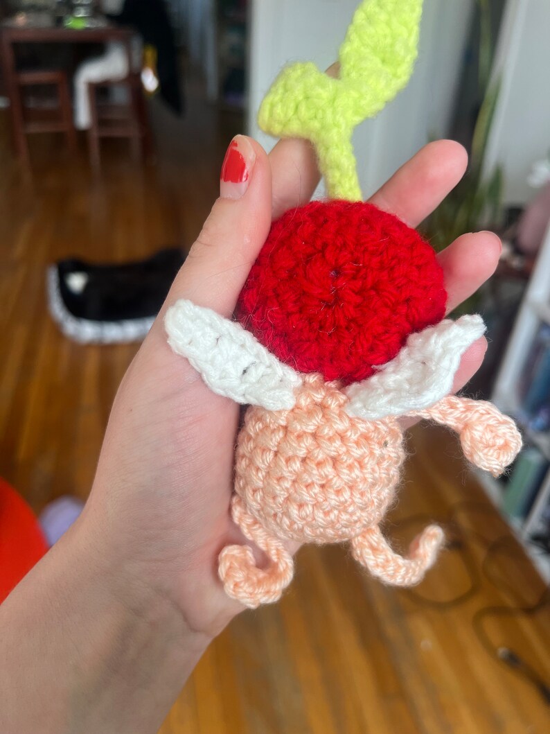 May include: A crocheted plush toy shaped like a cherry with a green stem and white wings. The cherry is red and the body is peach.