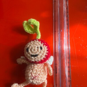 May include: A crocheted amigurumi toy shaped like a person with a red and pink body, green hair, and white wings. The toy is about 10 inches tall and is sitting on a red surface.