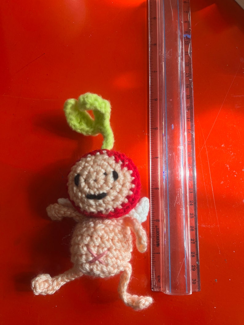 May include: A crocheted amigurumi toy shaped like a person with a red and pink body, green hair, and white wings. The toy is about 10 inches tall and is sitting on a red surface.