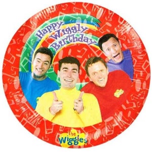 May include: Red paper plates with a colorful design featuring the four members of The Wiggles. The plates have a rainbow design with the words "Happy Wiggly Birthday" and "The Wiggles" printed on them.
