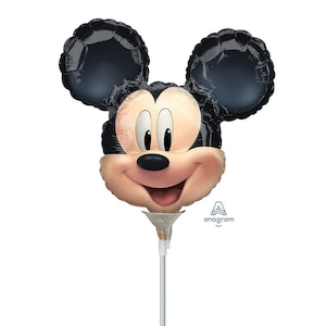 May include: A Mickey Mouse-shaped balloon with a smiling face, black ears, and a light tan face. The balloon is attached to a white stick with a silver-colored cone. The Anagram logo is visible on the right side.