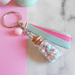 May include: A keychain featuring a small glass bottle filled with colourful marshmallows, attached to a silver ring and a pink and blue ribbon.