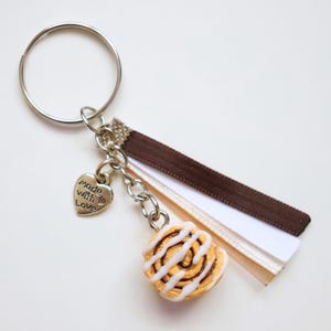 May include: A silver keyring with a miniature cinnamon roll charm, a heart-shaped charm that says "made with love", and a brown and white ribbon. The cinnamon roll is light brown with white icing. The keyring is on a white background.