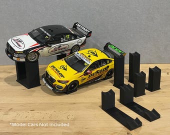 May include: A collection of black plastic display stands designed for model cars. The stands feature a vertical support with a curved cradle and a horizontal base. Two race car models are shown resting on two of the stands for demonstration.