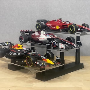 Könnte beinhalten: Drei Formel-1-Rennwagen-Modelle auf einem mehrstufigen Präsentationsständer. Die Autos sind rot, schwarz und weiß, mit Sponsorenlogos. Die Präsentation hat eine schwarze Basis und transparente Acrylregale.
