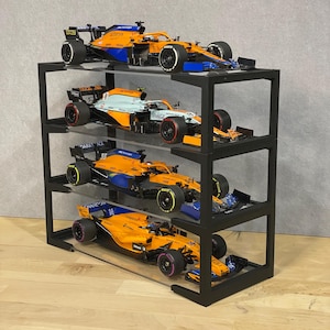May include: Four orange and blue Formula 1 race cars are displayed on a black metal shelf unit with clear acrylic shelves. The cars are arranged in a vertical stack, with the top car being the highest. The cars are all in different positions, with some facing forward and others facing sideways. The cars are all in different scales, with some being larger than others.