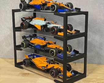 May include: Four orange and blue Formula 1 race cars are displayed on a black metal shelf unit with clear acrylic shelves. The cars are arranged in a vertical stack, with the top car being the highest. The cars are all in different positions, with some facing forward and others facing sideways. The cars are all in different scales, with some being larger than others.