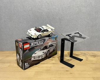 May include: A white Lego Speed Champions Lamborghini Countach set with a black stand. The set is on a wooden surface. The box has the text "LEGO SPEED CHAMPIONS" on it.