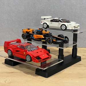 Puede incluir: Un expositor de tres niveles con tres coches de juguete. La plataforma inferior presenta un coche deportivo rojo. La plataforma central tiene un coche de carreras negro y naranja. La plataforma superior muestra un coche deportivo blanco. El soporte es negro con estantes de acrílico transparente.