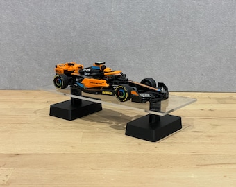 May include: A detailed model of a Formula 1 race car, predominantly orange and black, with sponsor logos. The car is displayed on a clear acrylic stand with black supports, set against a neutral background.