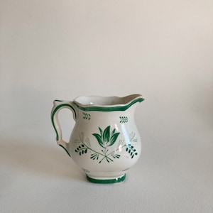 May include: A white ceramic creamer with a green floral design. The creamer has a scalloped rim and a handle.