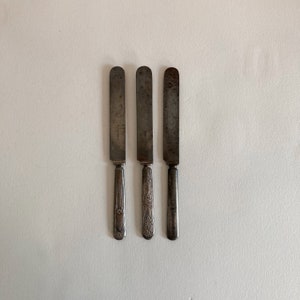 May include: Three silver butter knives with ornate handles. The knives are arranged in a row on a white surface.