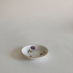 May include: A small white ceramic dish with a gold rim and a floral design featuring purple and yellow flowers.