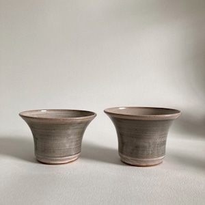 May include: Two handmade ceramic cups with a flared shape and a neutral color palette. The cups have a light gray exterior with a textured, wood-grain-like pattern and a lighter rim. The base is a lighter shade of brown. These cups are ideal for beverages.