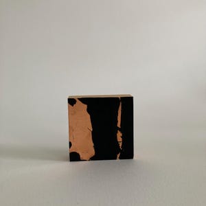May include: A small, square wooden block with a natural wood top and sides. The front and side surfaces are painted with a black, abstract design over a copper-colored base. The block is displayed against a white background.