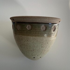 May include: A handmade ceramic wall planter with a speckled, light green and tan exterior. The top rim is a darker brown, and the planter features small, raised floral designs. This unique piece is ideal for displaying small plants or succulents.
