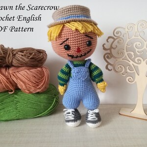 May include: A crocheted scarecrow doll with a blue and green striped shirt, blue overalls, and a brown and blue hat. The doll is standing on a table with yarn balls and a wooden tree cutout.