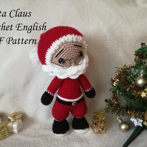 Puede incluir: Muñeco de Papá Noel de ganchillo con un traje rojo, barba blanca y botas negras. El muñeco está de pie frente a un pequeño árbol de Navidad con adornos dorados.