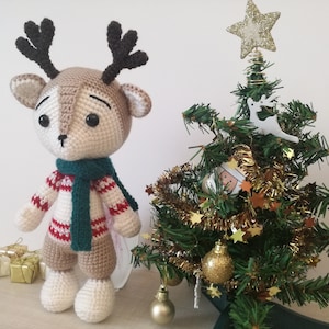 May include: A crocheted reindeer toy wearing a green scarf and a red and white striped jumper stands in front of a small artificial Christmas tree decorated with gold baubles and tinsel.