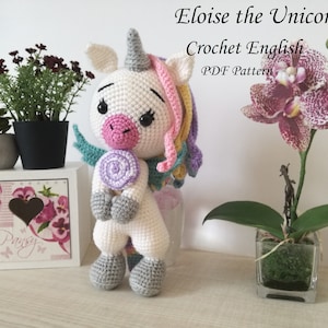 May include: A crocheted white unicorn toy with a pink nose, gray hooves, and a rainbow mane and tail. The unicorn is holding a purple and white swirl lollipop. The toy is sitting on a white shelf with a floral design.