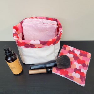Peut inclure: Un panier de rangement en tissu blanc avec une doublure à imprimé cœur rose et rouge. Le panier est rempli de disques démaquillants roses. Un pinceau à maquillage, une bouteille de liquide et un crayon à maquillage sont sur une surface noire à côté du panier.