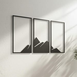 Op de afbeelding: Een set van drie zwarte metalen wandkunstwerken. Elk stuk heeft een rechthoekig frame met een berg silhouet ontwerp aan de onderkant. De kunst is gemonteerd op een witte, getextureerde muur, wat een moderne uitstraling creëert.