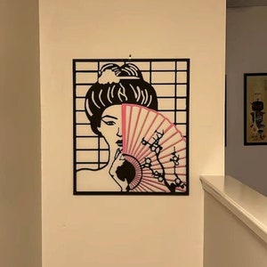 May include: Black metal wall art featuring a woman's face partially obscured by a pink and white fan with cherry blossoms. The artwork is set against a grid background.