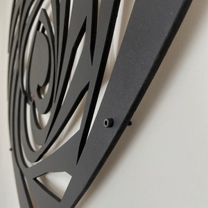 May include: Black metal wall art with a geometric design. The artwork features a spiral and triangular cut-out pattern. The piece is mounted on a white wall, showcasing the contrast between the dark metal and the light background.