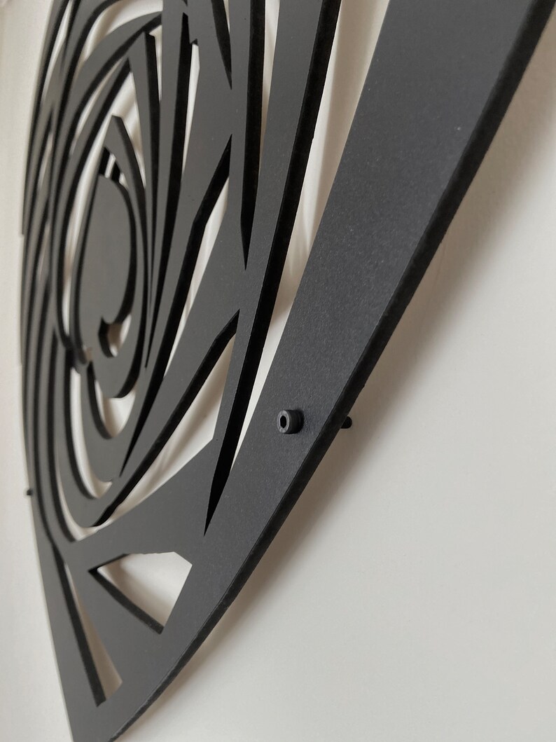 May include: Black metal wall art with a geometric design. The artwork features a spiral and triangular cut-out pattern. The piece is mounted on a white wall, showcasing the contrast between the dark metal and the light background.