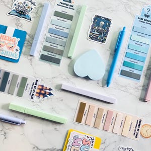 May include: A collection of stationery items including several sets of rectangular sticky note tabs with printed text, various pastel-colored pens and highlighters, a heart-shaped sticky note pad, and multiple decorative stickers with illustrations.